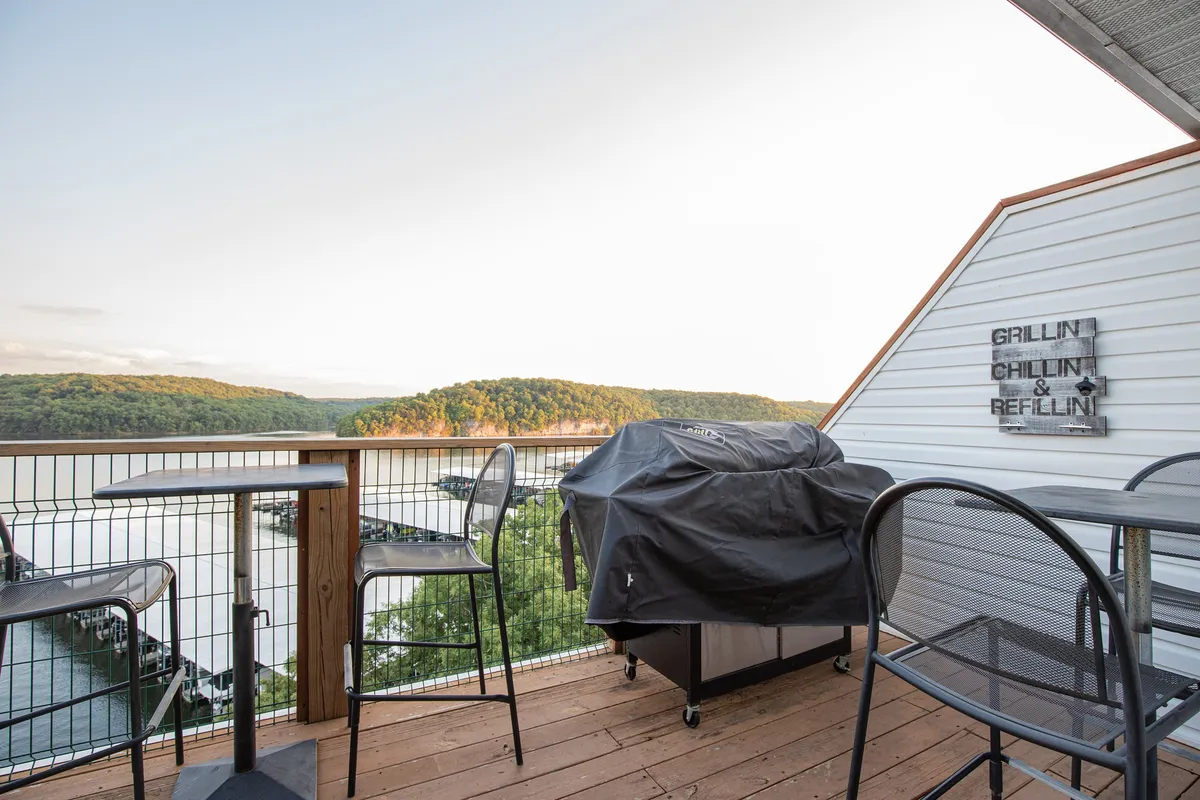 A beautifully arranged balcony with a grill set up, cozy outdoor seating, and an uninterrupted lake view stretching beyond the railing. The sign with text adds a welcoming touch, and lush greenery frames the vista, emphasizing the connection to nature.