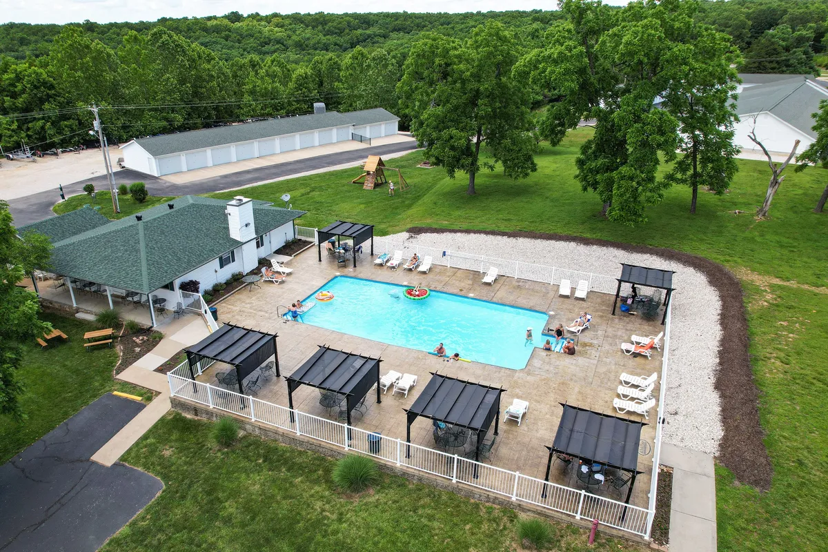 A lively exterior view of a swimming pool surrounded by covered seating areas, green trees, and playground equipment. People can be seen swimming, socializing, and children playing, creating a dynamic atmosphere.