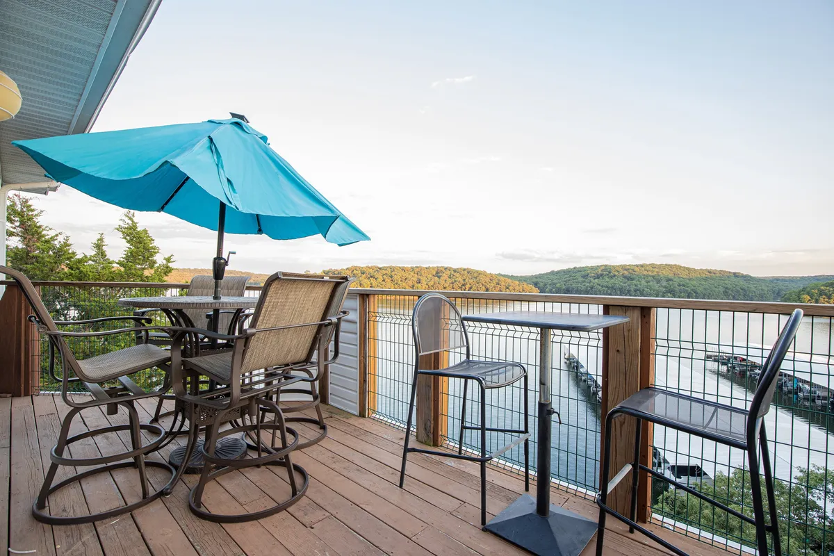 The image showcases a spacious wooden deck outfitted with a striking blue umbrella, overlooking a tranquil lake framed by lush trees. Inviting outdoor seating offers a perfect spot for dining, relaxing, or socializing, all set against a serene natural backdrop.