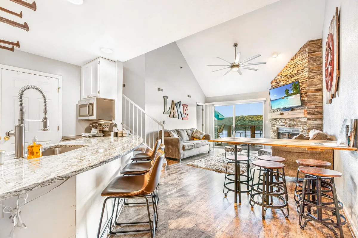 A bright, open living area featuring a comfortable sofa set against large windows that flood the space with natural light. A decorative stone wall and a wall-mounted TV create a cozy entertainment hub. The granite countertop hints at a modern kitchen just beyond the living zone, inviting conversations and group activities. The sunset view visible through the windows enhances the welcoming atmosphere.
