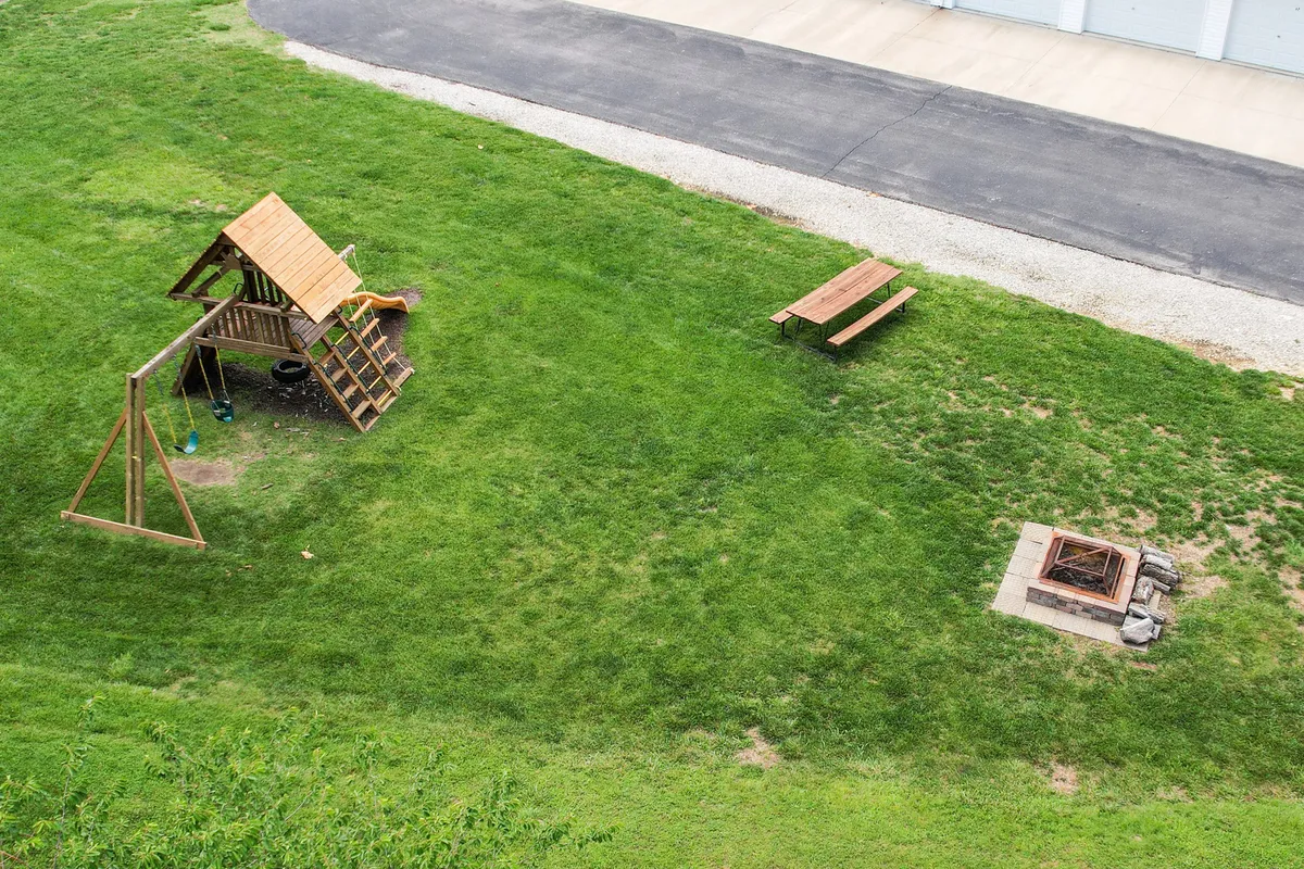 An exterior scene showcasing a playground structure, a picnic table, and a fire pit, all surrounded by trees and open space. The environment is inviting for families, children, and social gatherings, emphasizing outdoor recreation.