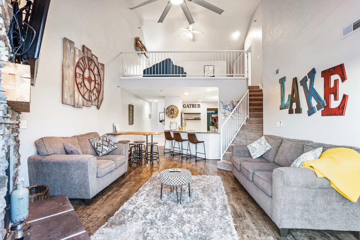 A welcoming living space featuring a comfortable sofa, coffee table, lake-themed decor, a 'gather' sign, and an inviting staircase. The scene suggests a space designed for relaxing with family, entertaining guests, or enjoying cozy evenings by the fireplace.