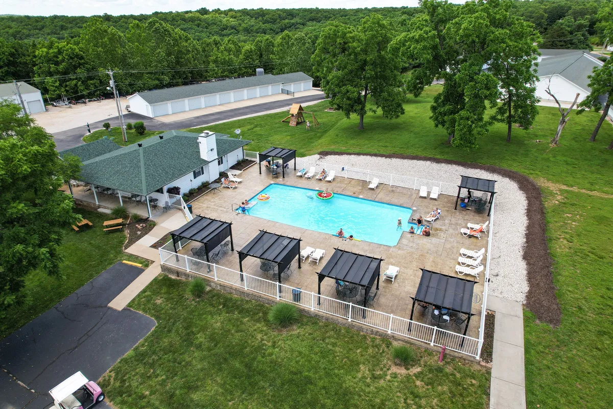 This image displays an exterior resort pool area with cabana structures, a green lawn, and a play area where guests can relax, swim, and socialize in a cheerful, lively setting. Children are seen playing, and the overall atmosphere is energetic and communal.