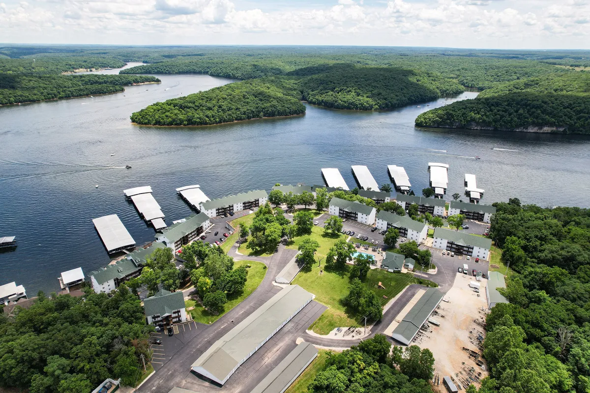 A sweeping exterior shot featuring the lake, boat docks, lush greenery, a swimming pool, and residential buildings. The image captures both the water and the adjacent park-like grounds, suggesting immediate access to natural adventures and relaxation.