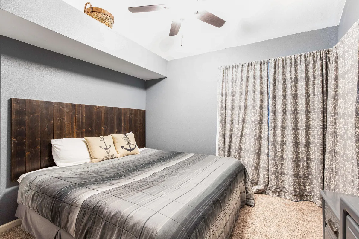 A serene king bedroom featuring a wooden headboard, decorative pillows, soft curtains, and an inviting bed with crisp linens. Natural light filters through the window, highlighting the restful atmosphere and the sense of spaciousness provided by high ceilings.