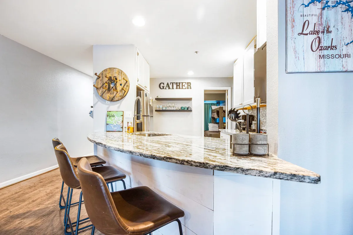 A welcoming bar area with granite countertops and comfortable bar stools anchors the gathering space. The prominent 'GATHER' sign and decorative wall art set a warm, communal tone, while the open view to surrounding rooms enhances the sense of connectivity and shared experiences.