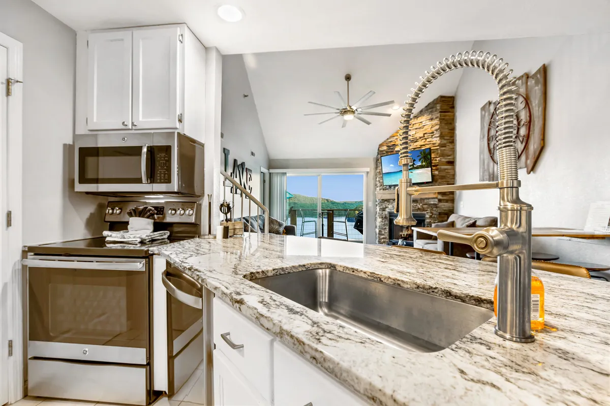 A modern, open kitchen with granite countertops, stainless steel appliances, a stone fireplace nearby, and large windows letting in ample light. The space is arranged for cooking, dining, and entertaining, with a seamless flow between kitchen and living areas.