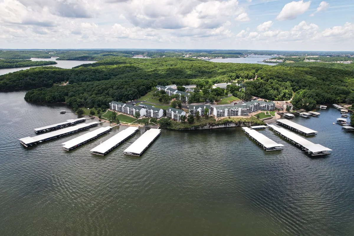 A wide view of the lake and marina, with residential buildings nestled among greenery and a tranquil atmosphere. The scene captures the allure of lakeside living and the opportunities for boating or relaxing by the water, all framed by Missouri's natural beauty.